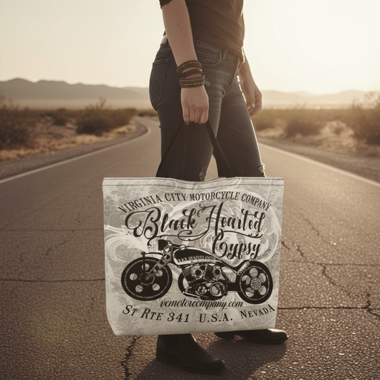VCMCo. Black Hearted Gypsy canvas tote bag held at hip level on a Nevada desert road at golden hour — close-up lifestyle shot showing bold chopper and script graphic, biker lifestyle apparel