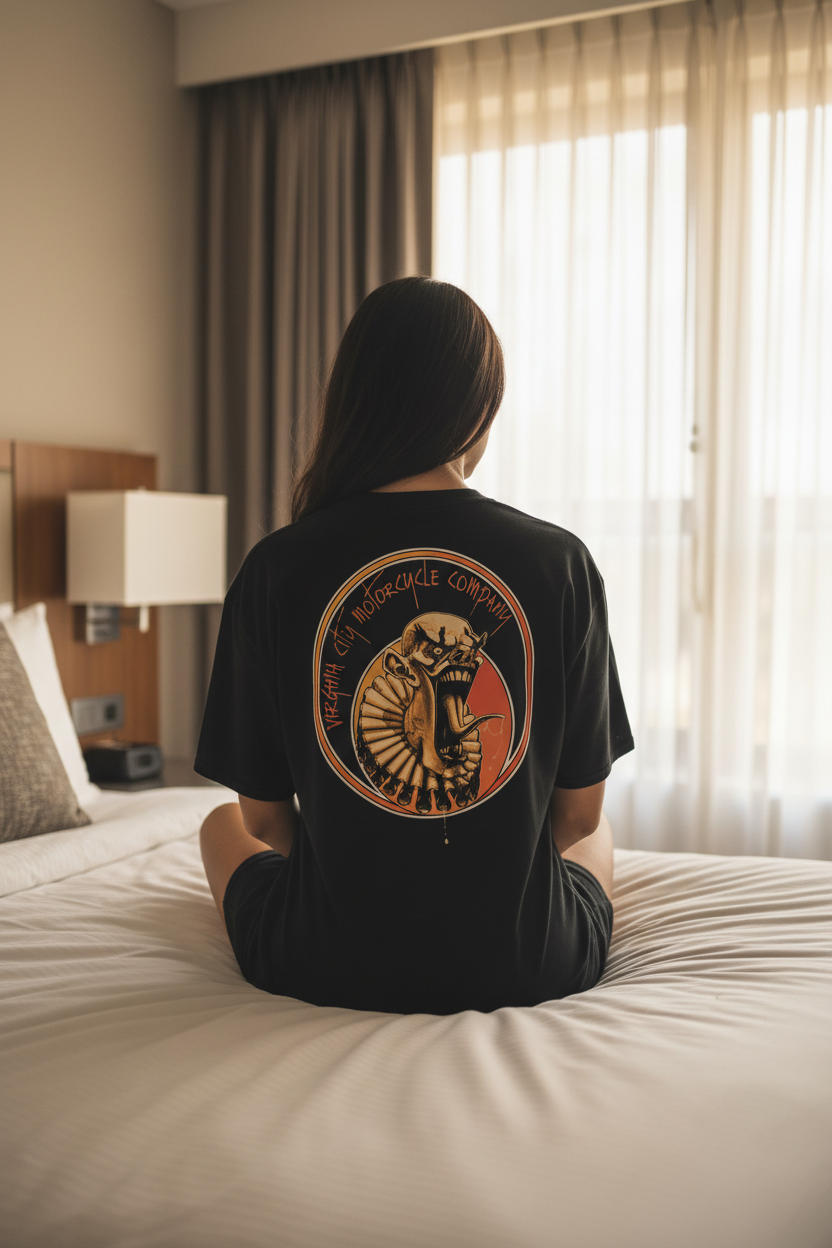 Clown Series #2 Black Biker T-Shirt back graphic — woman seated on hotel room bed wearing oversized tee, bold orange and yellow clown artwork by Virginia City Motorcycle Company, VCMCo. Men's motorcycle graphic tee.