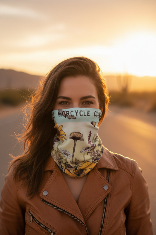 Close-up of a woman rider wearing the VCMCo. Rustic Wildflowers Neck Gaiter pulled up over her nose and chin, featuring a botanical wildflower print with yellow sunflowers, purple lavender, white daisies, and butterflies on a soft sky-blue and cream background, with a golden Nevada desert highway at golden hour behind her.