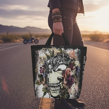 VCMCo. Metal Head and Flowers canvas tote bag held at hip level on a Nevada desert road at golden hour — close-up lifestyle shot showing crowned skull and floral graphic, biker lifestyle apparel