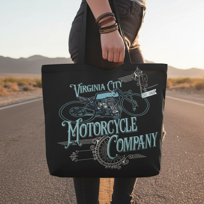 VCMCo. Victorian Bike canvas tote bag held at hip level on a Nevada desert road at golden hour — close-up lifestyle shot showing teal and silver ornate motorcycle graphic, biker lifestyle apparel