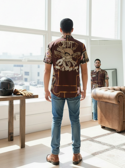 Man in blue jeans and riding boots stands back-to-camera in a sun-drenched urban loft, showcasing the Wrenchin' Since 1969 skull and crossed wrenches graphic on the back of the VCMCo. dark brown chain-print moto dress shirt, with mirror reflection revealing the shirt front — helmet and leather gloves on table nearby.